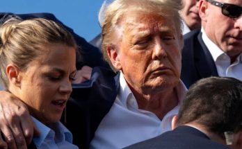 Donald Trump with eyes closed and blood on his face being supported by Secret Service