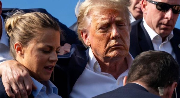 Donald Trump with eyes closed and blood on his face being supported by Secret Service