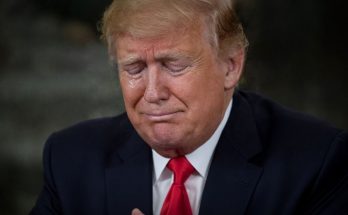 Donald Trump with eyes closed and a tear on his cheek, wearing a dark suit and red tie