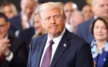 Donald Trump in a dark suit and red tie standing before a blurred crowd, with text above