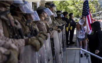 A line of law enforcement officers in riot gear with shields faces a woman holding a
