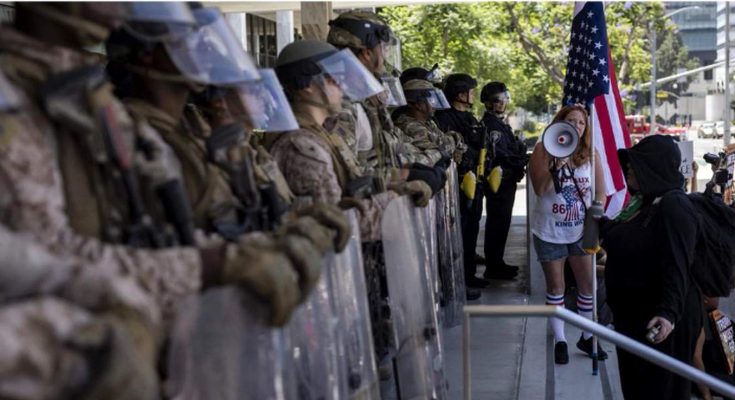 A line of law enforcement officers in riot gear with shields faces a woman holding a