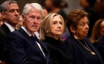 Bill Clinton and Hillary Clinton seated together at a formal event, both wearing dark