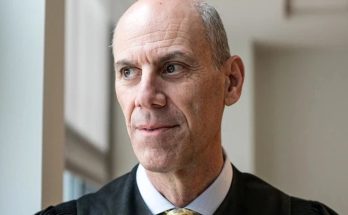 A bald man with gray hair on the sides wearing a black judicial robe with a white collar