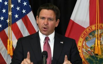 Ron DeSantis speaking at a podium with American and Florida state flags behind him, with