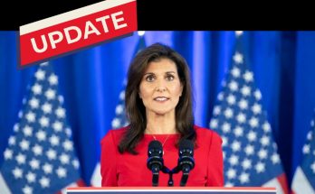Nikki Haley in a red dress standing at a podium with two microphones, American flags