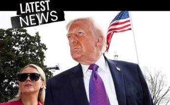 Donald Trump in a dark suit and purple tie stands outdoors with an American flag behind