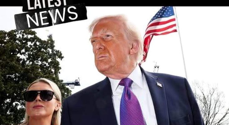 Donald Trump in a dark suit and purple tie stands outdoors with an American flag behind