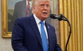 Donald Trump speaking at a podium with a microphone, wearing a dark blue suit and light