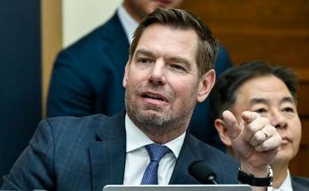 A man in a dark blue suit with a blue patterned tie sits at a table with a microphone