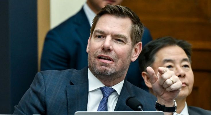 A man in a dark blue suit with a blue patterned tie sits at a table with a microphone
