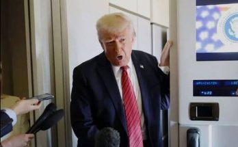 Donald Trump in a dark suit and red striped tie speaking to reporters with microphones