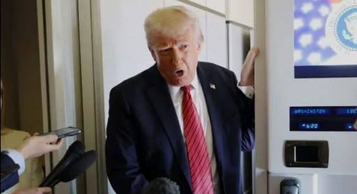 Donald Trump in a dark suit and red striped tie speaking to reporters with microphones