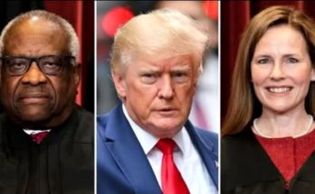 Donald Trump in a dark suit and red striped tie speaking to reporters with microphones
