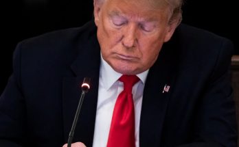 Donald Trump in dark suit, white shirt, and red tie, looking down with eyes closed