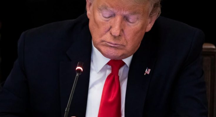 Donald Trump in dark suit, white shirt, and red tie, looking down with eyes closed