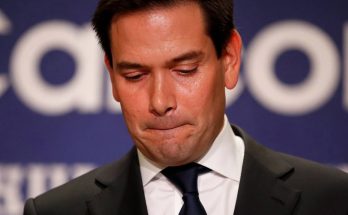 A man in a dark suit and blue tie looks downward with a somber expression, standing