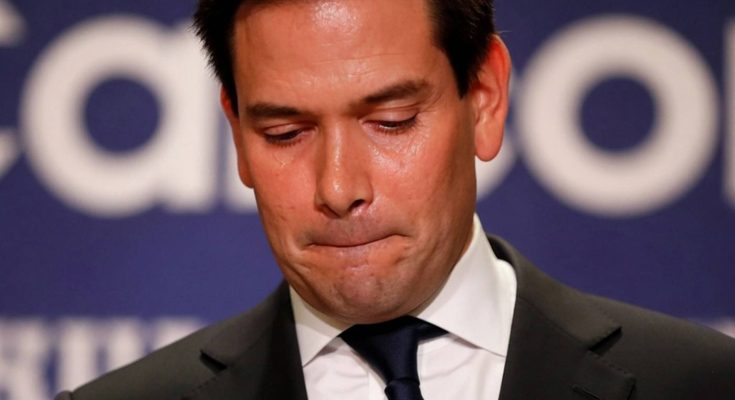 A man in a dark suit and blue tie looks downward with a somber expression, standing