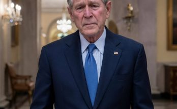 George W. Bush in a navy suit and blue tie standing in an ornate hallway with chandeliers
