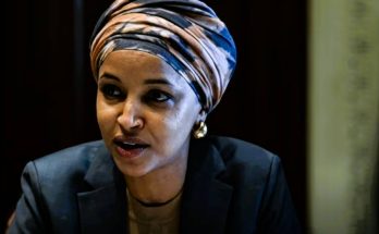 A woman wearing a striped turban and dark blazer speaks while looking to her right