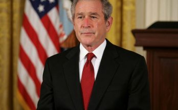 George W. Bush in a black suit and red tie standing in front of an American flag and