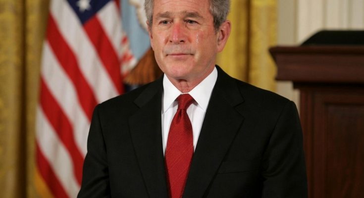 George W. Bush in a black suit and red tie standing in front of an American flag and