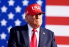 Donald Trump wearing a red USA cap and red tie stands in front of an American flag with