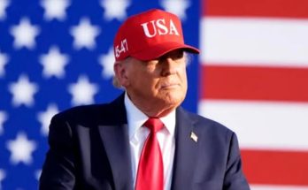 Donald Trump wearing a red USA cap and red tie stands in front of an American flag with