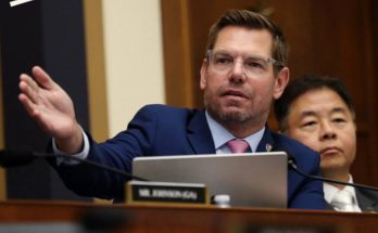 A man in a blue suit and pink tie sits at a desk with a microphone and laptop, gesturing