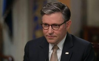 A man with gray-streaked dark hair wearing glasses, a dark suit, white shirt, and tan