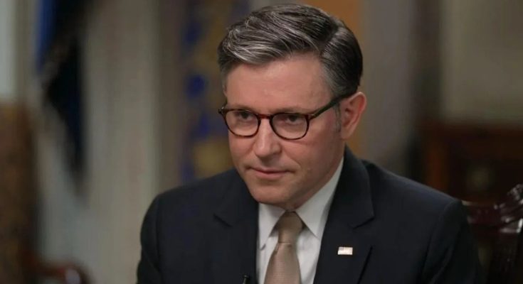 A man with gray-streaked dark hair wearing glasses, a dark suit, white shirt, and tan