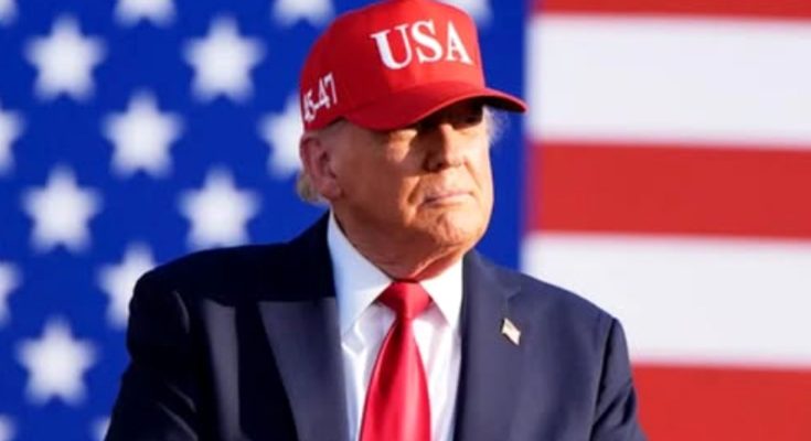 Donald Trump wearing a red USA cap and red tie stands in front of an American flag with