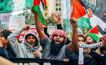 A crowd of protesters in an urban setting holding Palestinian flags and signs, with one