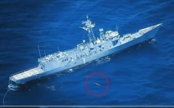 A gray naval frigate sailing on deep blue ocean water with a small object circled in red