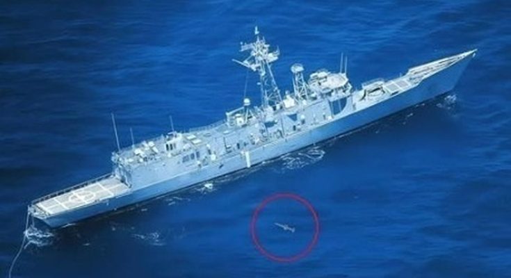 A gray naval frigate sailing on deep blue ocean water with a small object circled in red