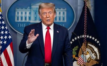 Donald Trump standing at a podium with microphones, wearing a dark blue suit, white