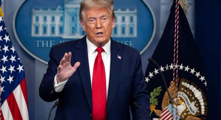 Donald Trump standing at a podium with microphones, wearing a dark blue suit, white