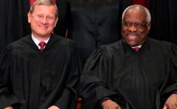 Two men in black judicial robes with red ties seated side by side, one with gray hair