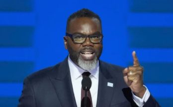 A man with a gray beard and glasses wearing a dark suit and tie speaks at a podium