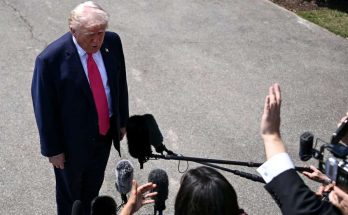 Donald Trump in a dark suit and red tie stands on pavement speaking to reporters holding