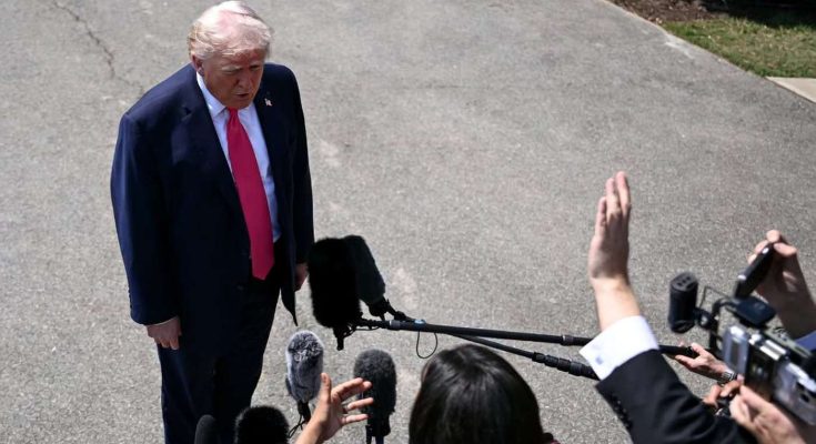 Donald Trump in a dark suit and red tie stands on pavement speaking to reporters holding