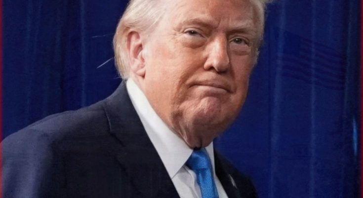 Man with blond hair wearing a dark suit and blue tie, standing in front of a blue curtain