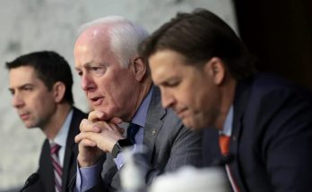 Three men in suits seated at a table with microphones, one speaking with hands clasped