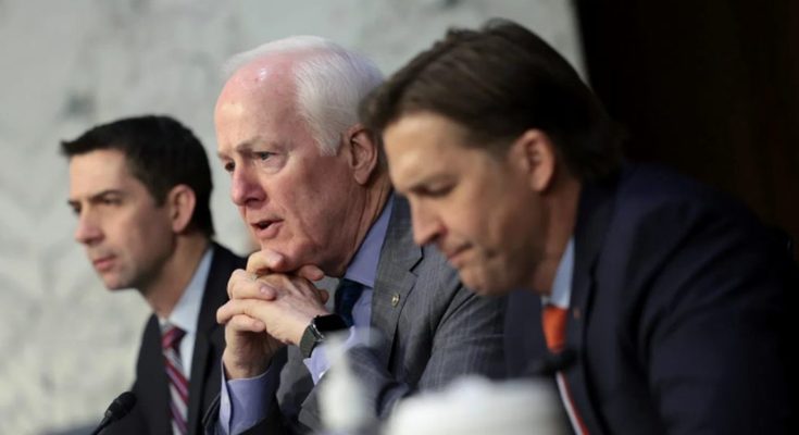 Three men in suits seated at a table with microphones, one speaking with hands clasped