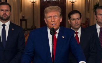 Donald Trump speaking at a podium with a microphone, wearing a blue suit and red tie