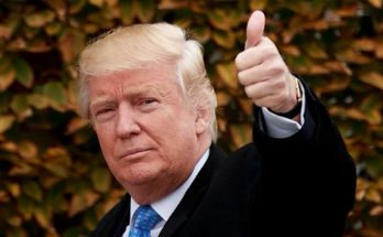 Donald Trump giving a thumbs up gesture with his right hand, wearing a black coat, white