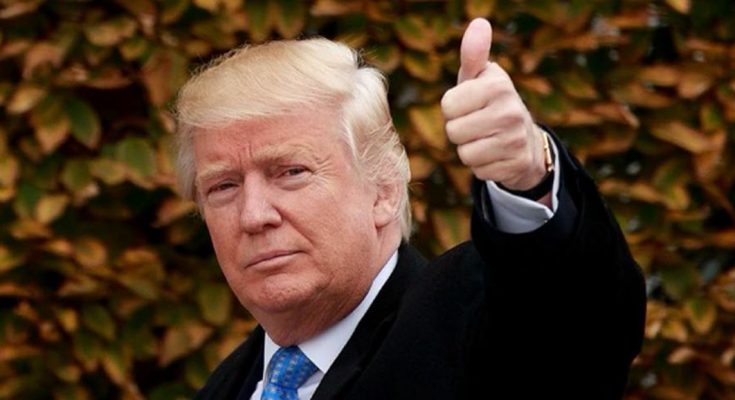 Donald Trump giving a thumbs up gesture with his right hand, wearing a black coat, white