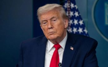 Donald Trump in a dark suit and red tie standing at a podium with a microphone, with an