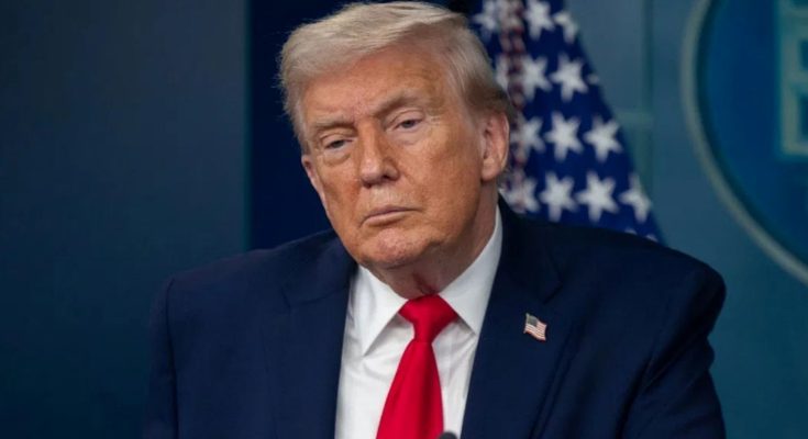 Donald Trump in a dark suit and red tie standing at a podium with a microphone, with an