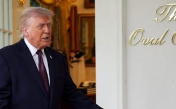 Donald Trump standing in a doorway with arms extended, wearing a dark suit and red tie